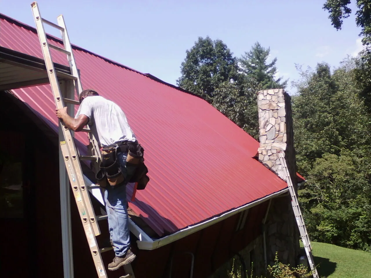 Roofer on a ladder beside a red metal roof and stone chimney — Soffit & Fascia Repair by Roofing Brothers in Knightdale